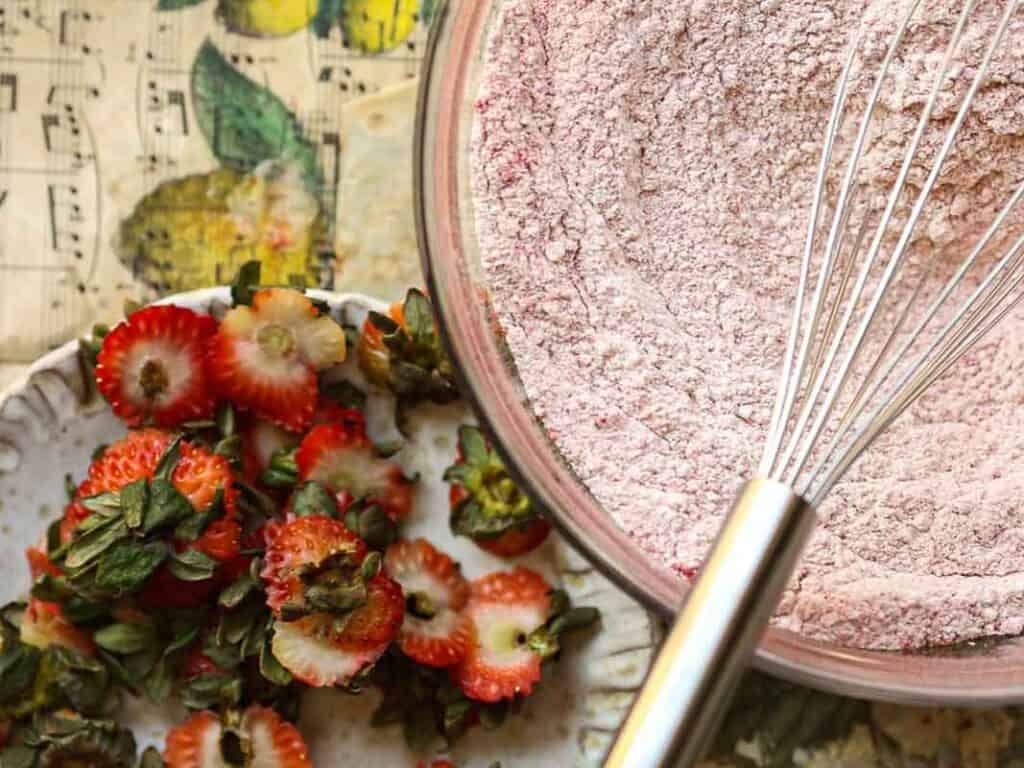 A close-up view of a mixing bowl filled with pinkish flour and a whisk. Beside the bowl, there are sliced strawberries with green tops on a plate. The background features a decorative music note pattern with yellow accents.